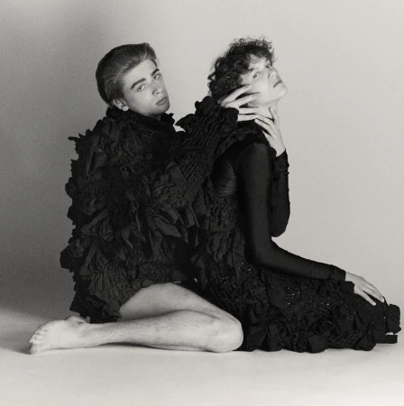 Two people pose in black clothing against a light background in a studio shot. One person is sitting with their hand touching the other person.