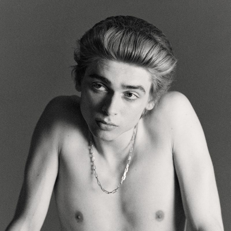 Black and white portrait of a young adult male with fair skin and light brown hair, wearing a silver chain necklace.