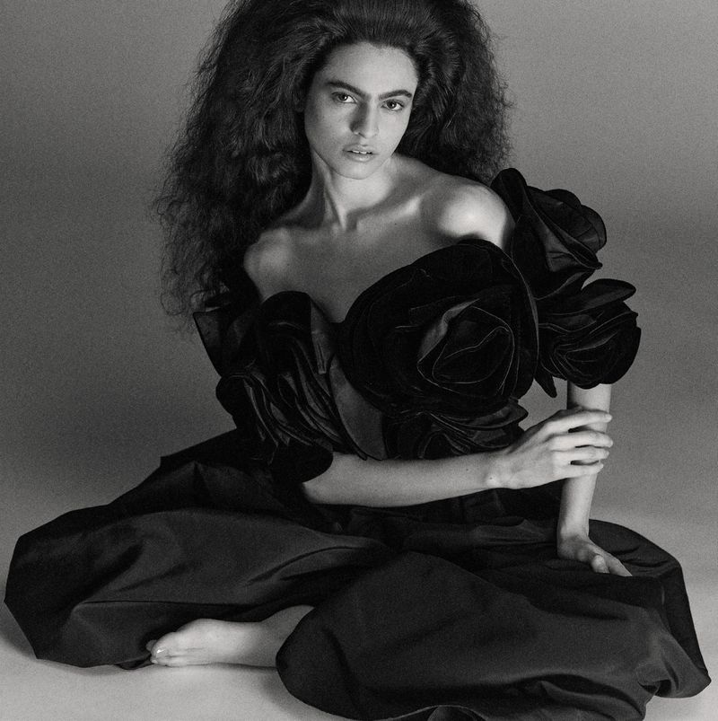 Black and white portrait of a woman in a dramatic dark dress with rose accents sitting in a studio with a serious expression.