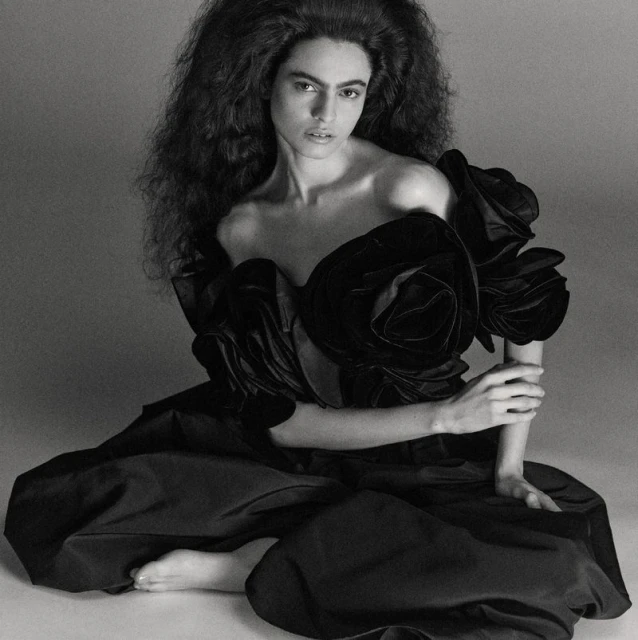 Black and white portrait of a woman in a dramatic dark dress with rose accents sitting in a studio with a serious expression.
