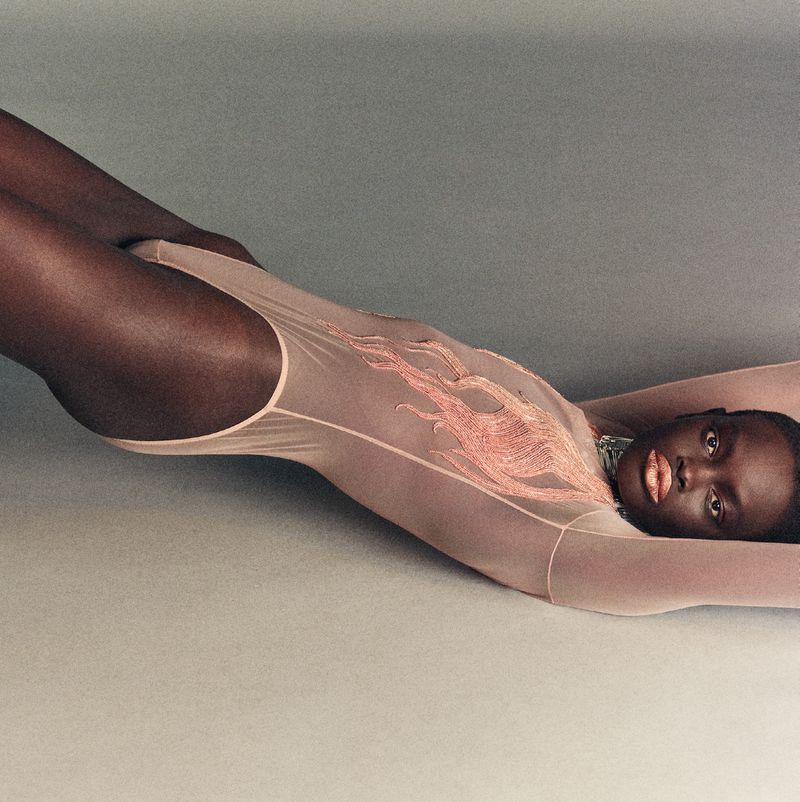 A dark-skinned woman in a light pink bodysuit with flame designs lies on her back with her arms raised in a studio setting.