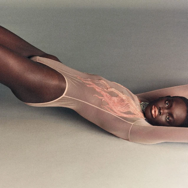 A dark-skinned woman in a light pink bodysuit with flame designs lies on her back with her arms raised in a studio setting.