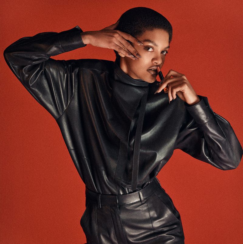 Close-up of an African-descent adult female in a black leather outfit, applying lipstick against a solid terracotta background in studio lighting.