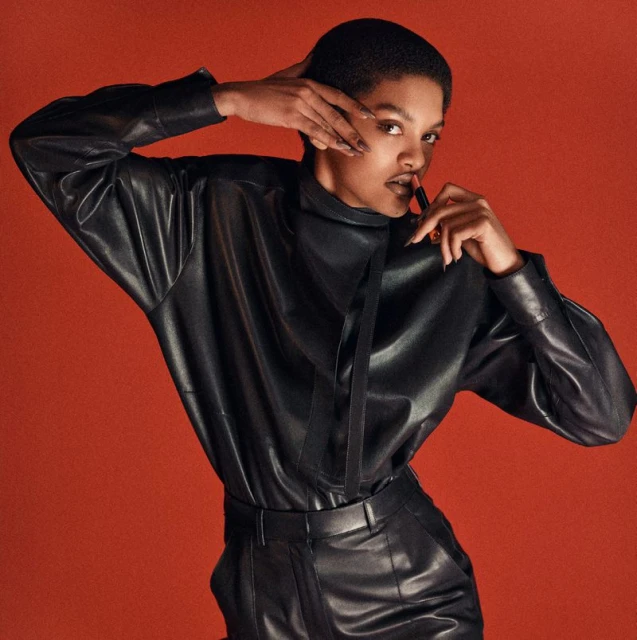 Close-up of an African-descent adult female in a black leather outfit, applying lipstick against a solid terracotta background in studio lighting.