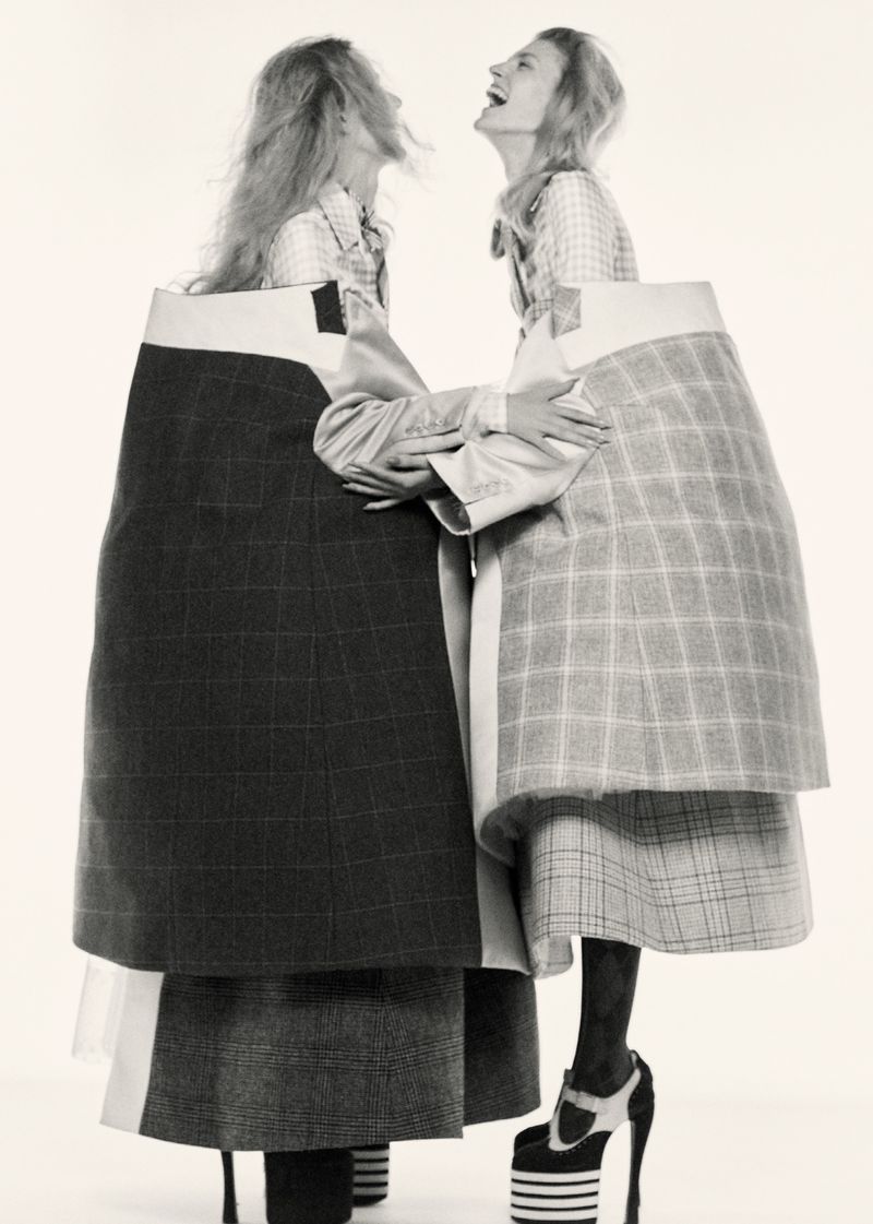 Two women in avant-garde fashion embrace against a white background, showcasing exaggerated plaid skirts and high platform heels.
