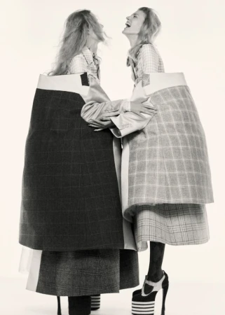 Two women in avant-garde fashion embrace against a white background, showcasing exaggerated plaid skirts and high platform heels.