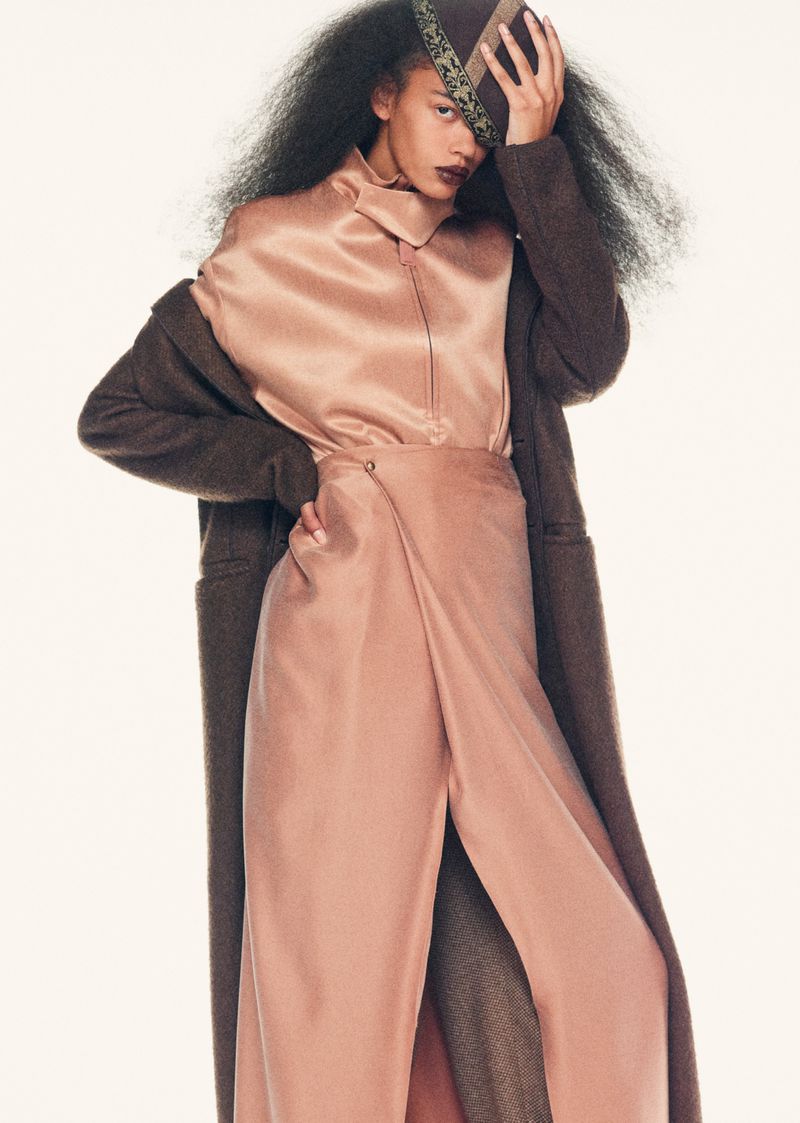 A young woman poses in a pink silk outfit, including a high-necked blouse and split-legged trousers, with a long brown coat.