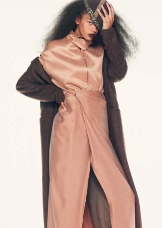A young woman poses in a pink silk outfit, including a high-necked blouse and split-legged trousers, with a long brown coat.