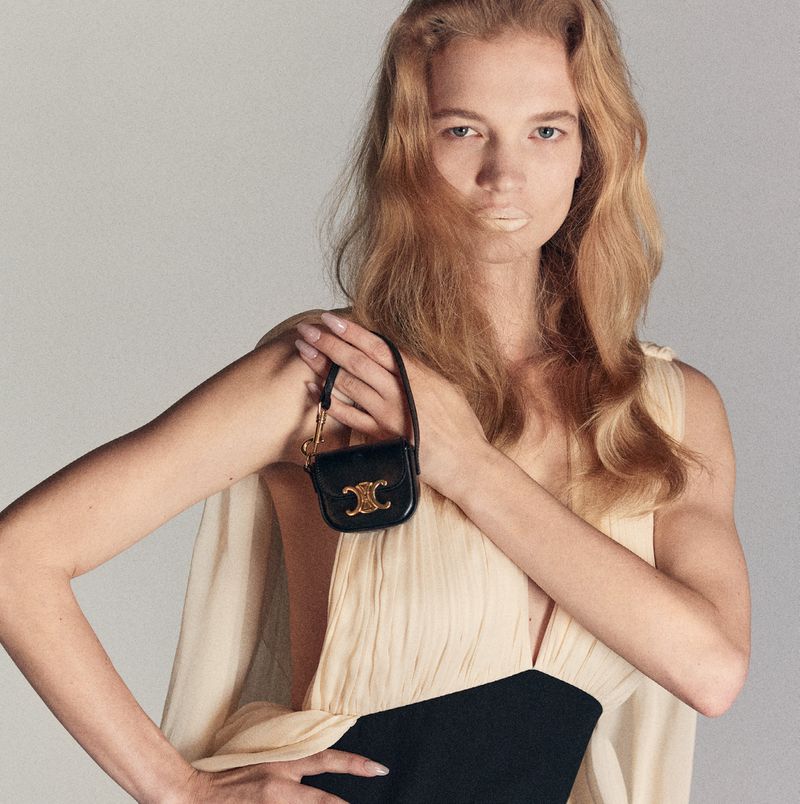 A light-skinned adult woman with long blonde hair is featured in a studio shot, holding a black leather purse with a gold-tone logo.