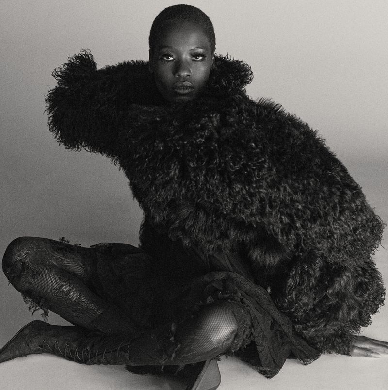 Monochrome portrait of an adult Black woman in a dramatic fur coat, seated in a relaxed pose in a light, neutral indoor studio setting.