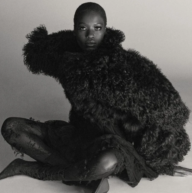 Monochrome portrait of an adult Black woman in a dramatic fur coat, seated in a relaxed pose in a light, neutral indoor studio setting.