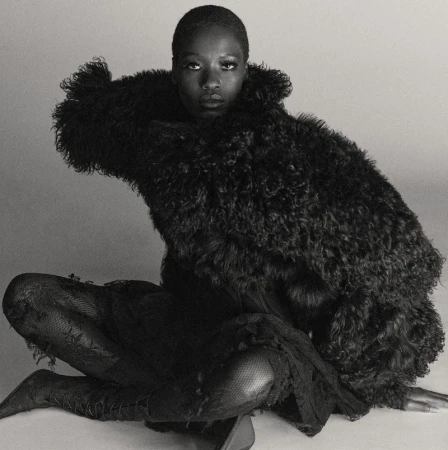 Monochrome portrait of an adult Black woman in a dramatic fur coat, seated in a relaxed pose in a light, neutral indoor studio setting.