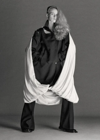 Model wearing dark structured jacket and trousers with white draped fabric against neutral studio background