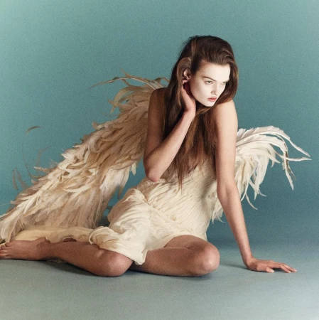 Model with large feathered wings wearing an off-white dress sitting against a muted teal background for System Magazine.