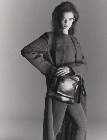 Monochrome fashion portrait of a model in a dark coat and trousers holding a leather shoulder bag against a grey backdrop.