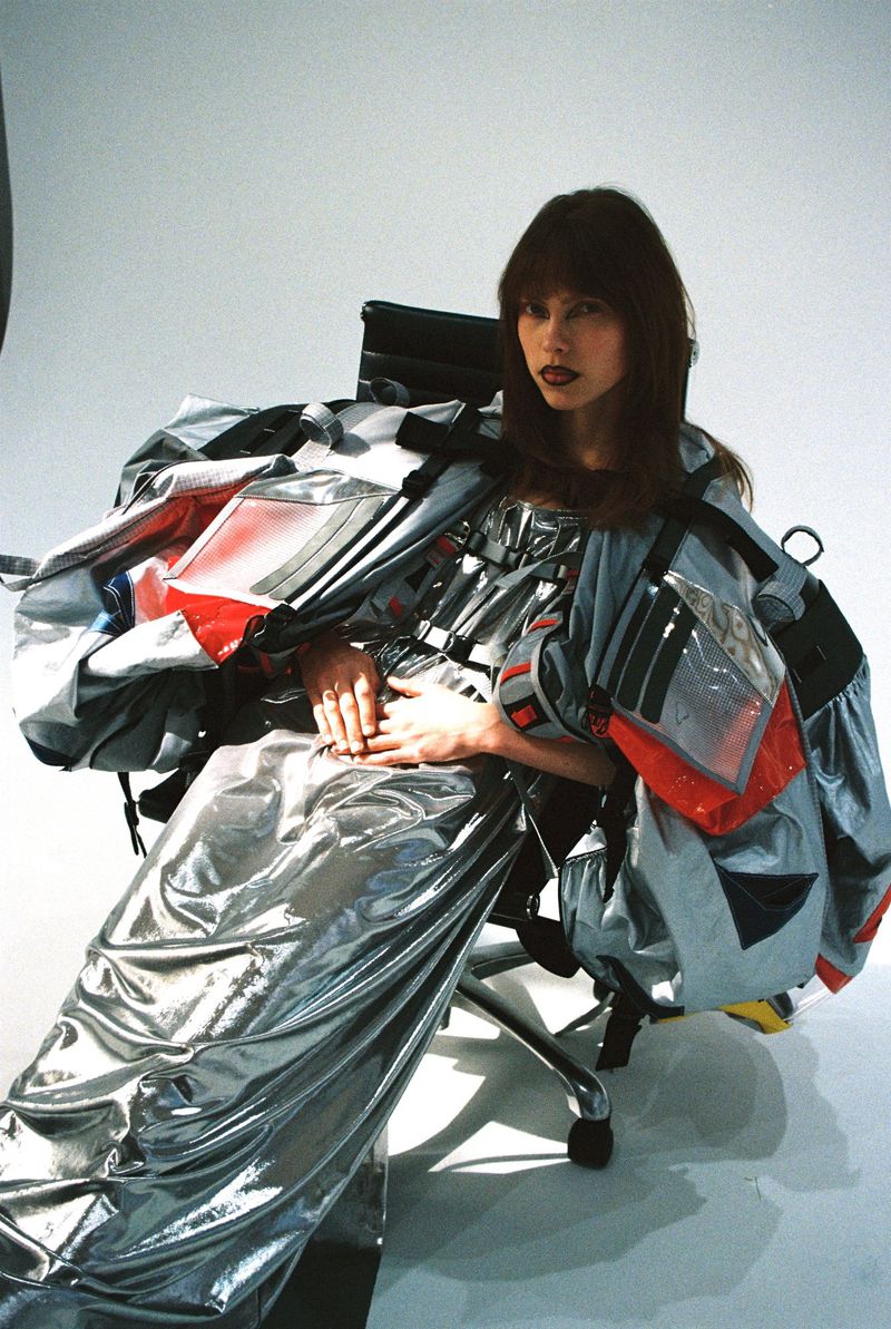 Woman in a futuristic silver gown sits in an office chair against a bright backdrop, showcasing unique fashion in a studio setting.