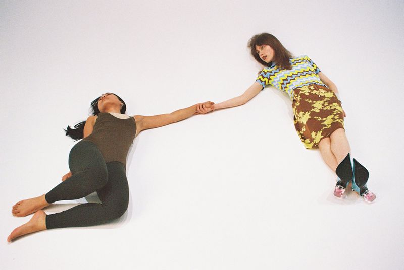 Two women lying on a white surface are holding hands, one in a brown top and leggings, and the other wearing a yellow patterned outfit.