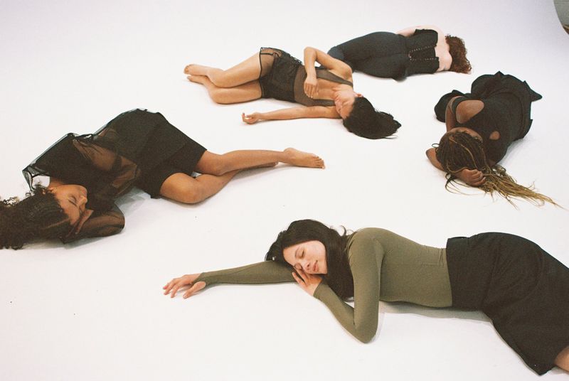 Six women of various ethnicities are lying down in a circle in a photo studio with white seamless background wearing dark-colored clothing.