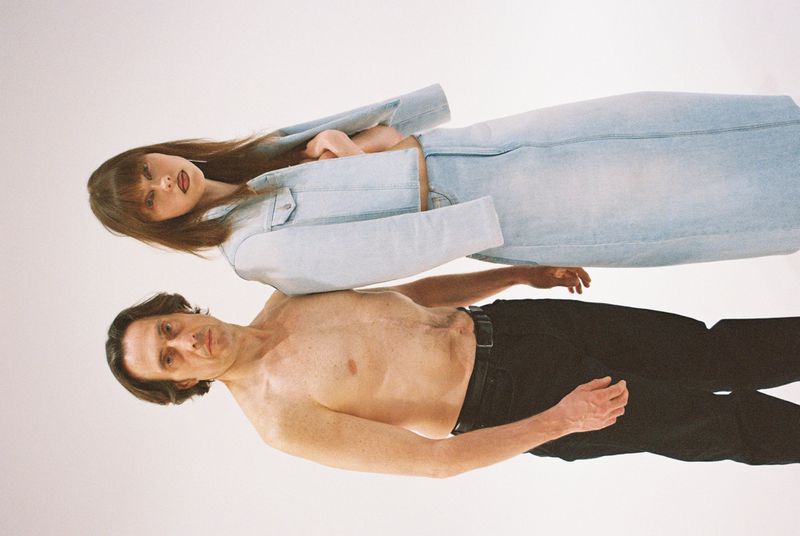 A woman in a denim jacket and skirt lies on top of a shirtless man against a white backdrop in a surreal, visually striking composition.