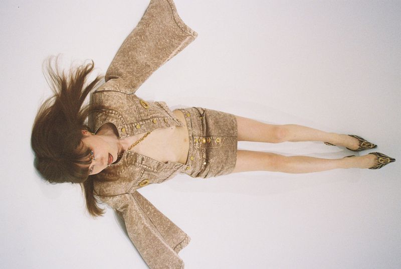 Fashion photo of a woman in a brown denim mini skirt and cropped jacket with gold buttons lying on a white surface, wearing high heels.