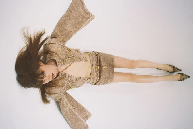 Fashion photo of a woman in a brown denim mini skirt and cropped jacket with gold buttons lying on a white surface, wearing high heels.