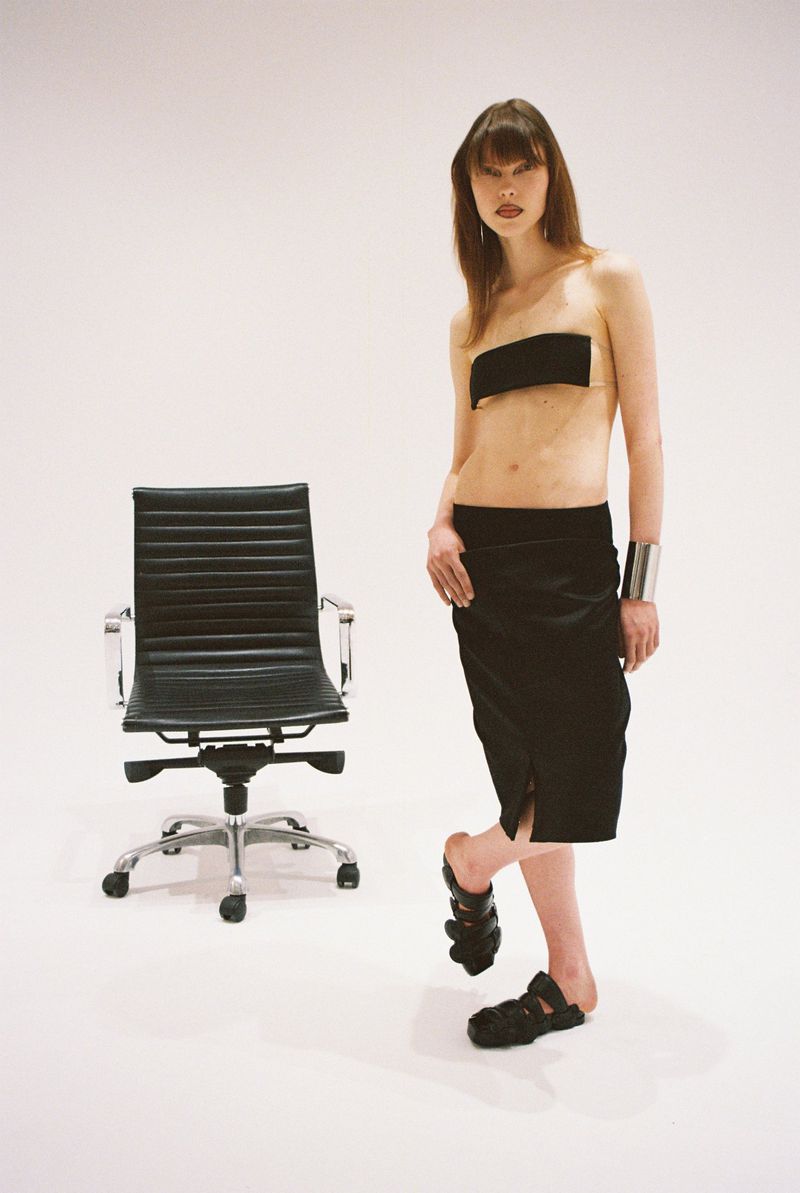 Fashion shot of a woman in a black outfit posing next to a black office chair against a white backdrop in a studio setting.