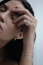 Close-up portrait of a woman with closed eyes holding her hand near her forehead, showcasing diamond rings and a bracelet.