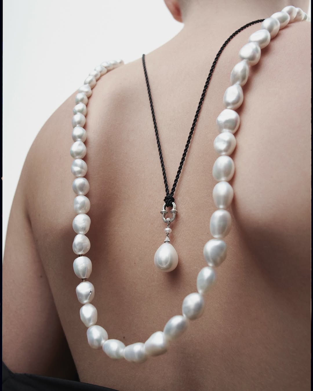 Back view of a person wearing a pearl necklace and a black cord necklace with a pearl pendant against a white background.