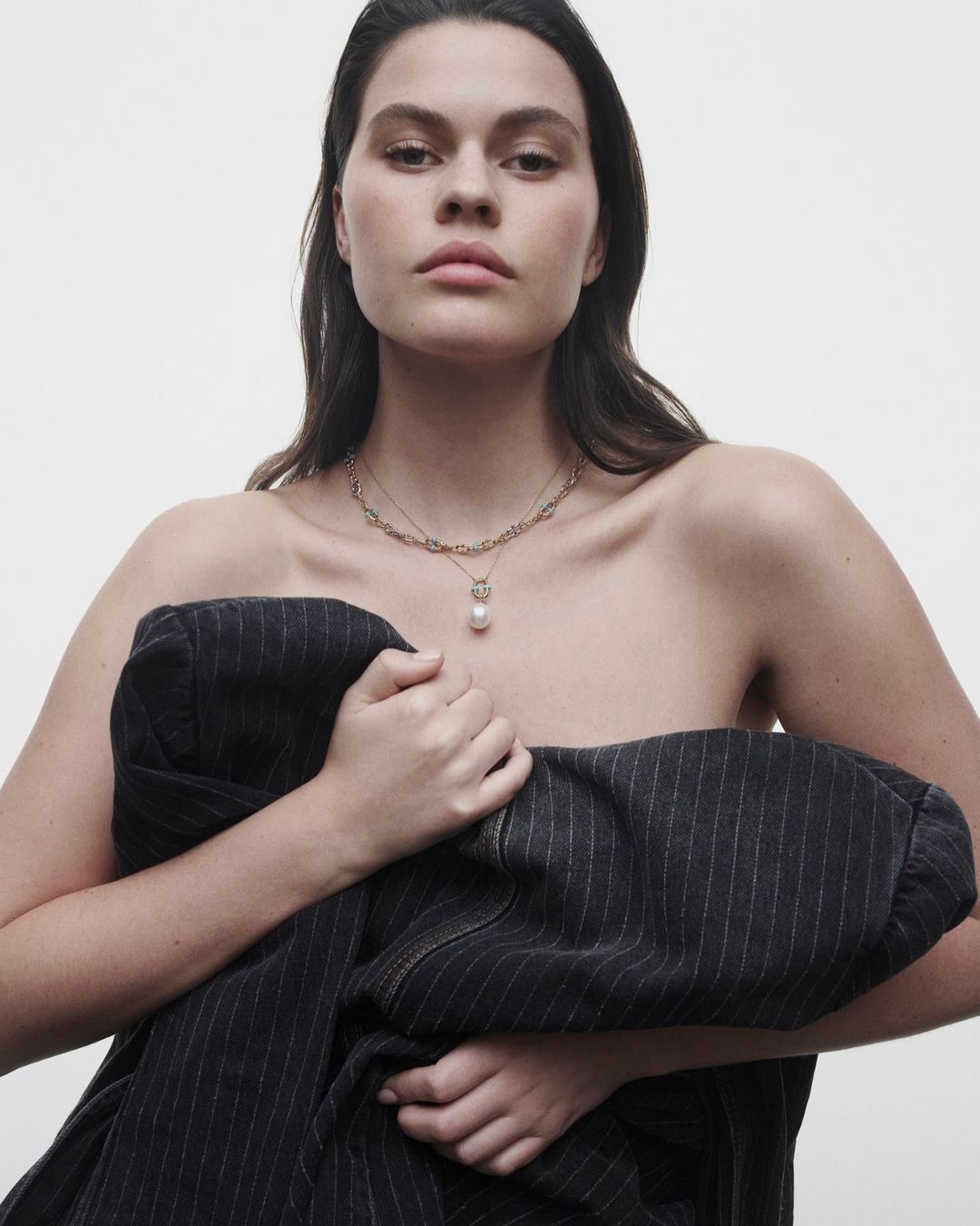 Striking beauty shot: Woman with brown hair wearing a gold necklace, draped in a pinstripe jacket against a bright background.