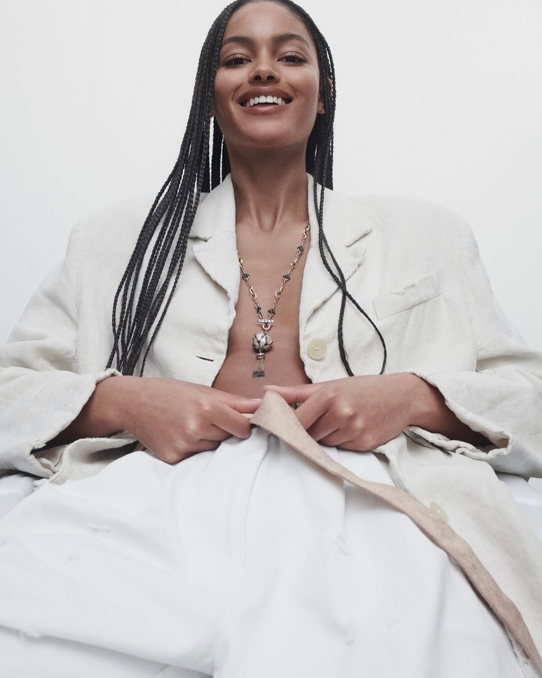 A smiling woman with braided hair wears a cream-colored blazer and necklace in a bright studio setting, angled from below in a fashion shot.