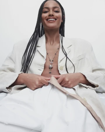 A smiling woman with braided hair wears a cream-colored blazer and necklace in a bright studio setting, angled from below in a fashion shot.