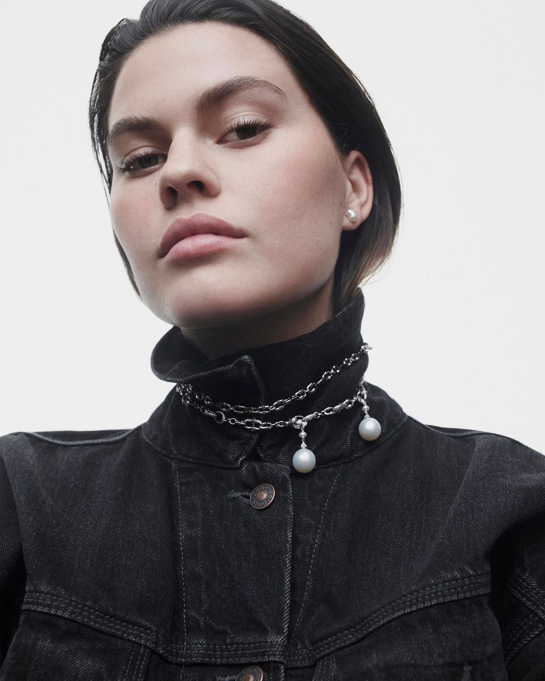Close-up of a woman wearing a black denim jacket and a pearl necklace. Studio shot on a white background for fashion or beauty advertising.