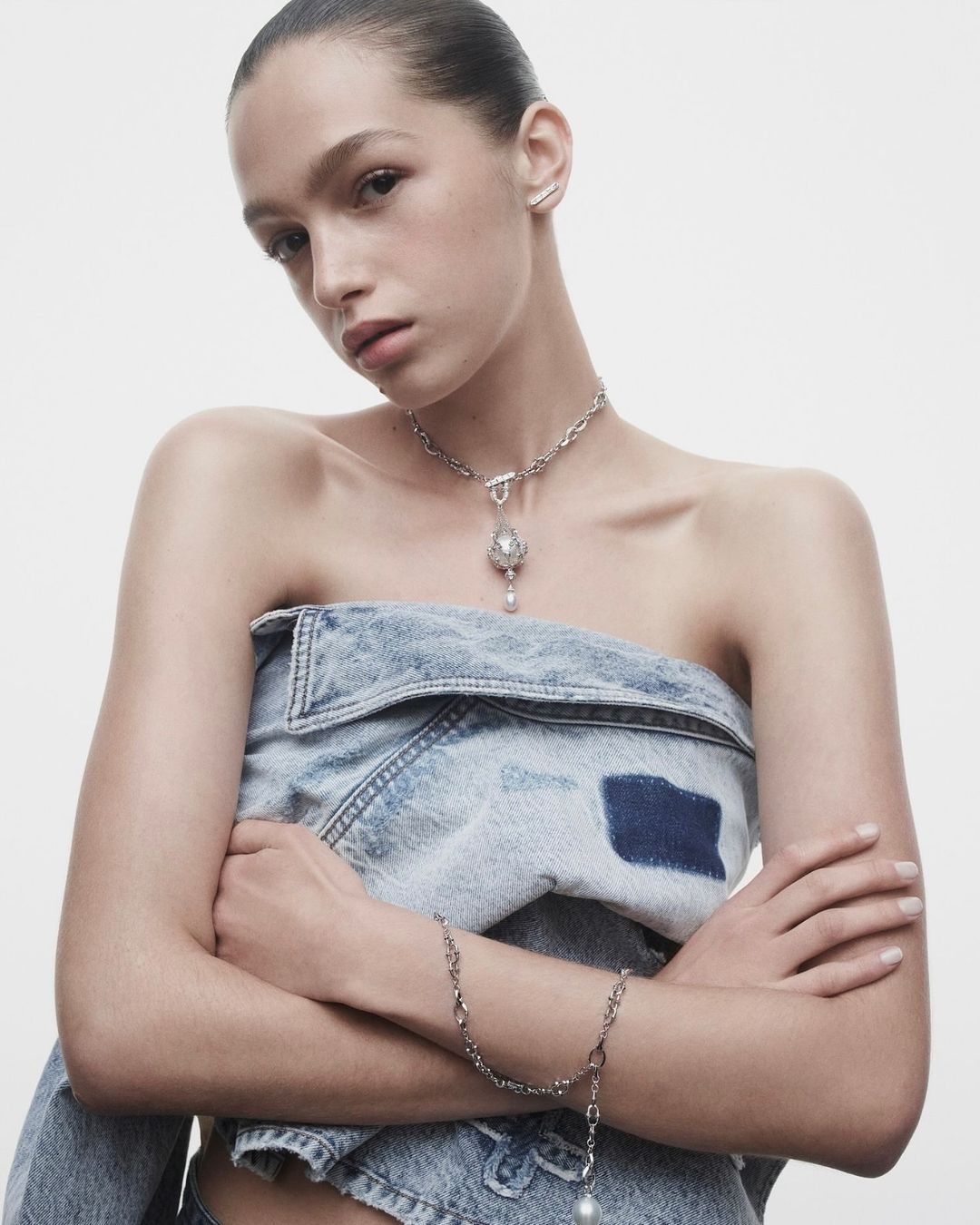Young woman poses in denim, wearing pearl jewelry in a bright studio shot, showcasing a bracelet and necklace.