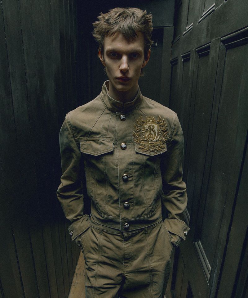 Fashion shot of a model in an olive-green jumpsuit with an ornate logo on the jacket, against a dark wood panelled wall.