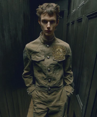 Fashion shot of a model in an olive-green jumpsuit with an ornate logo on the jacket, against a dark wood panelled wall.