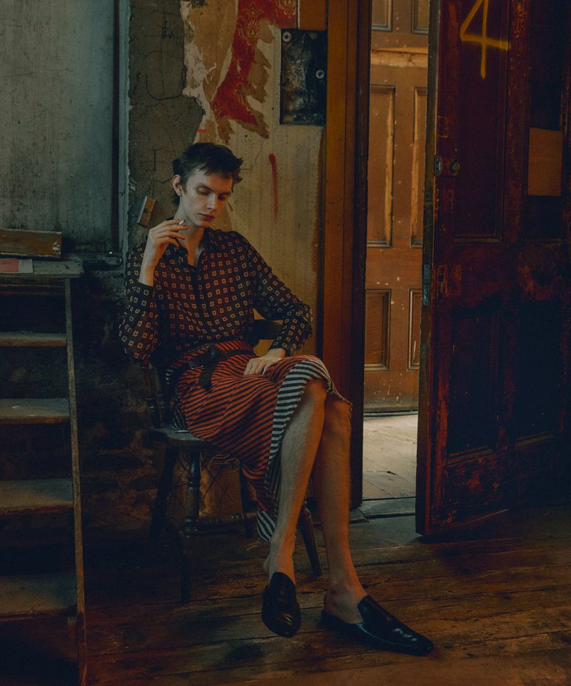 A slender man seated in a vintage doorway, wearing a patterned shirt and striped skirt, radiates a calm, poised presence in a dimly lit interior.