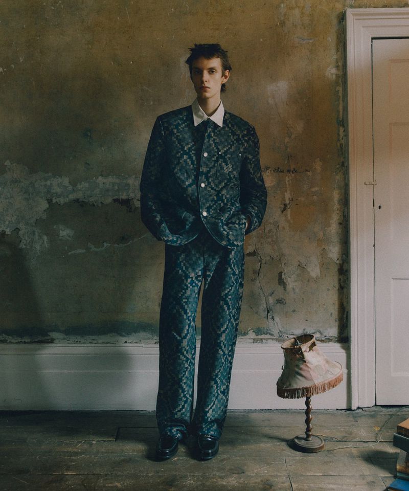 An adult male poses indoors wearing a patterned suit with a white collared shirt, dress shoes, and visible lamp and books.