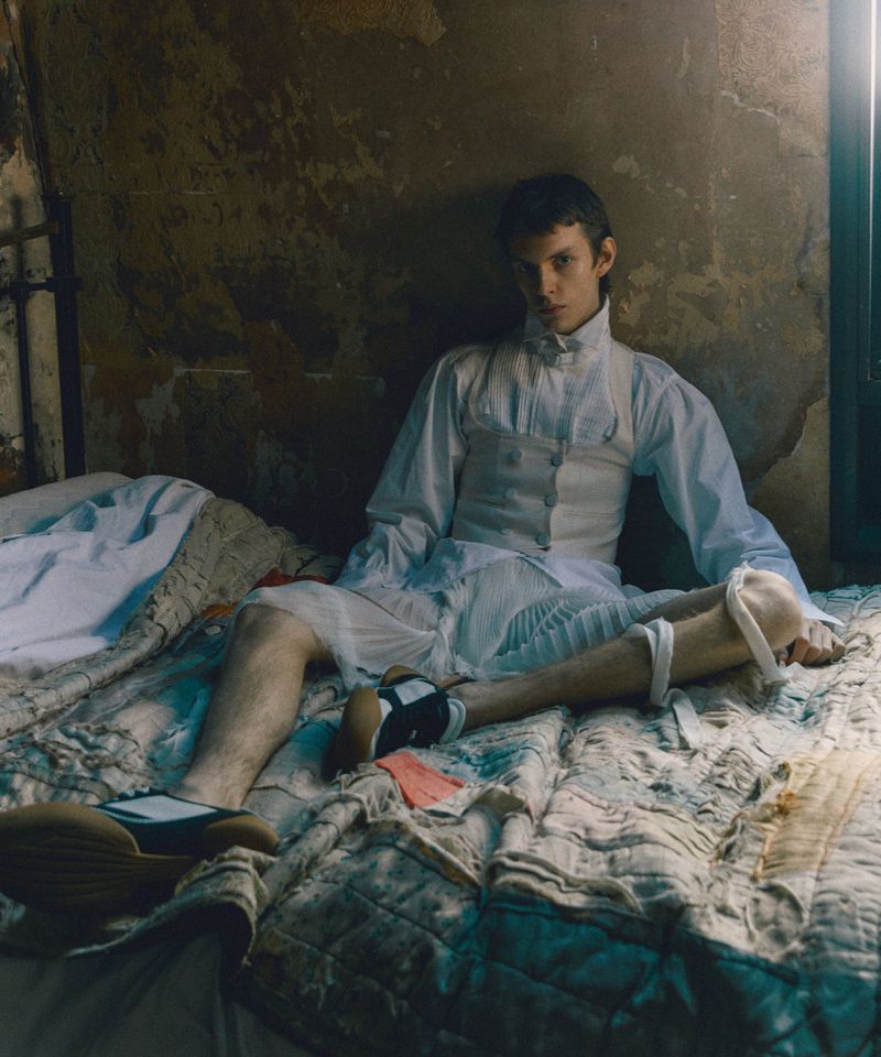 Man in period clothing sitting on a bed in a room with peeling paint, with muted colors and a serious expression on his face.