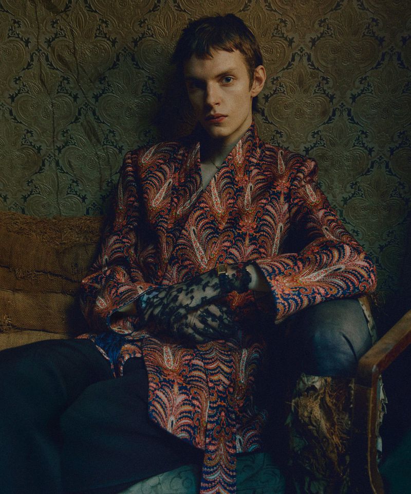 Portrait of a young adult man in a patterned robe sitting indoors. Close-up shows his expression, clothing detail, and interior setting.