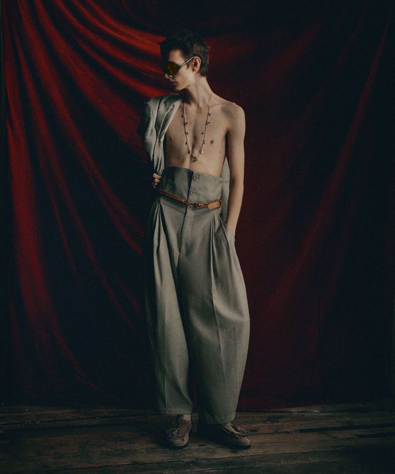 A fashion portrait of a slim man wearing oversized grey pleated trousers and a light grey jacket indoors against a red draped backdrop.
