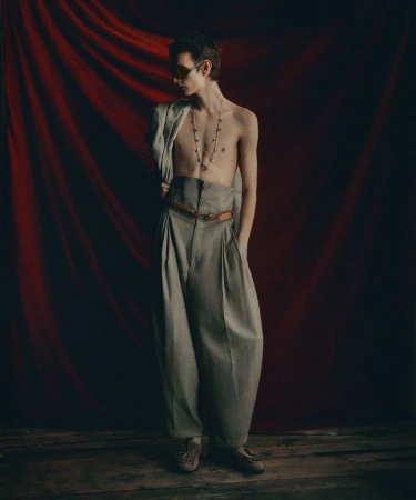 A fashion portrait of a slim man wearing oversized grey pleated trousers and a light grey jacket indoors against a red draped backdrop.