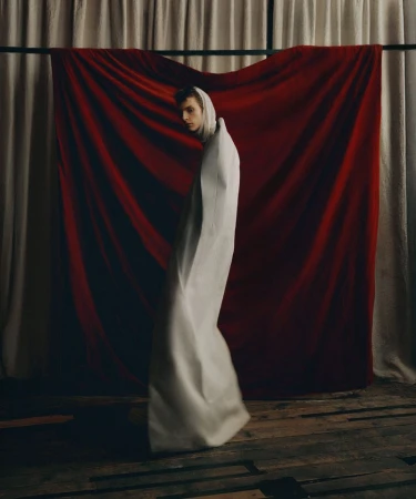 Figure standing, shrouded in white fabric, creating abstract shapes. Red velvet drapes in the background adds contrast to the composition.