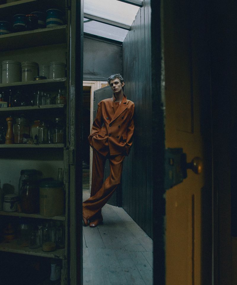 Fashion shot of a slender young adult in a bronze suit, posing indoors with jars and bottles on shelves visible in the background.