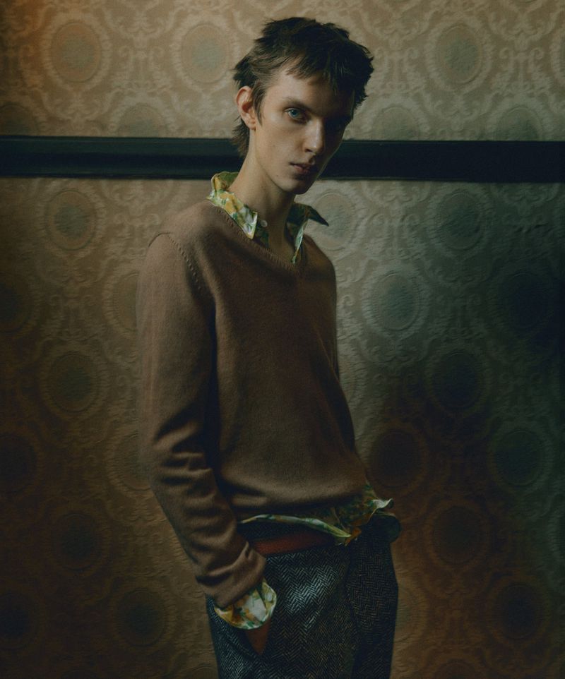 Portrait of a young adult male in casual fashion, wearing a brown v-neck sweater and patterned shirt, set against a patterned wall.