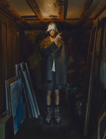 A fashion shot of a person in an overcoat and shorts, posing in an enclosed, dimly lit room with scattered canvases and wooden ceiling beams.