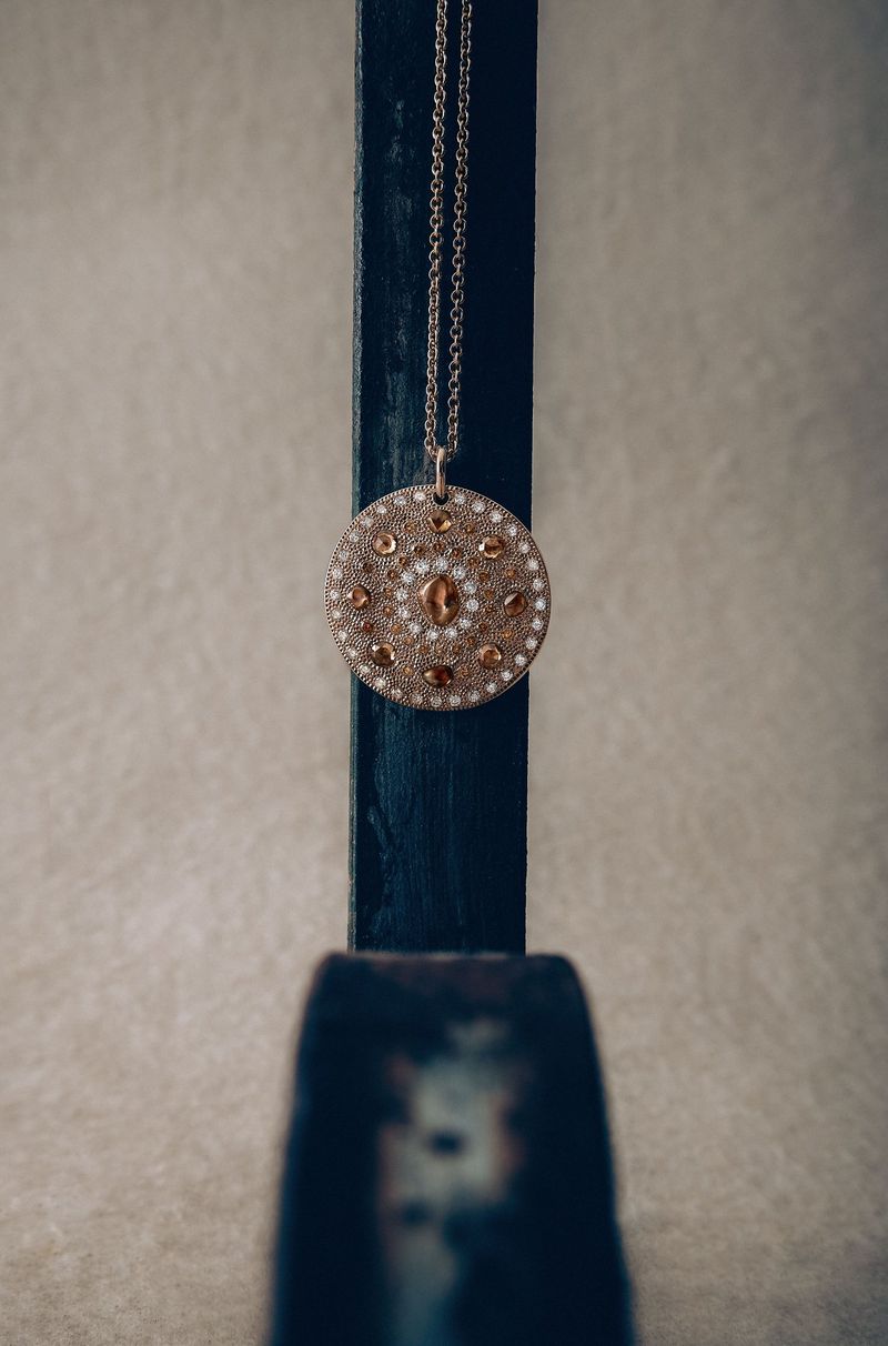 Close-up of a round pendant necklace with brown and white gems hanging from a dark blue wooden stand against a pale grey background.
