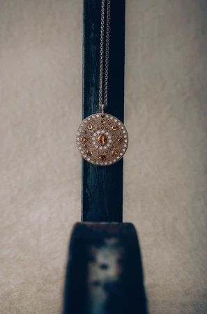 Close-up of a round pendant necklace with brown and white gems hanging from a dark blue wooden stand against a pale grey background.