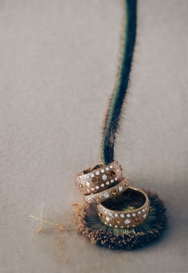 Close-up of three gold-toned, textured rings stacked on a green flower stem with a grey background in a vertical product shot.