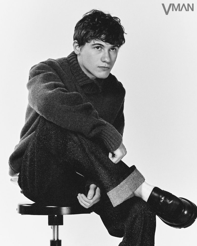 Black and white portrait of a young adult male seated on a stool. He wears a sweater and wool trousers, posing with a serious expression.