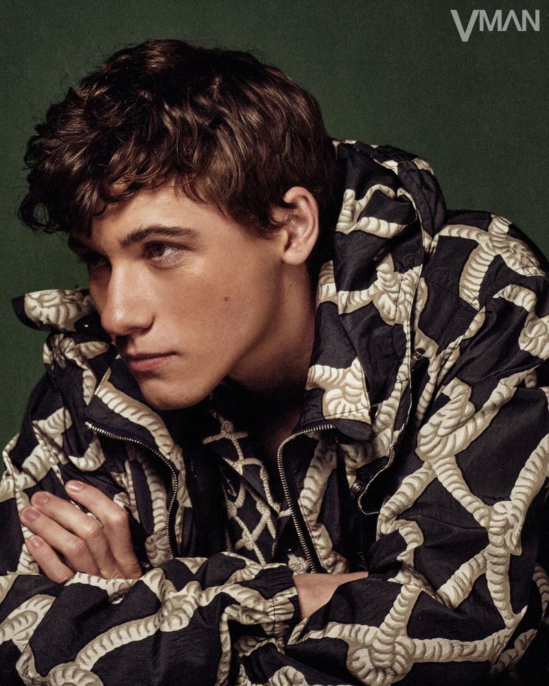 Portrait of a young man with dark, curly hair wearing a black jacket with white rope pattern against a dark green background indoors.